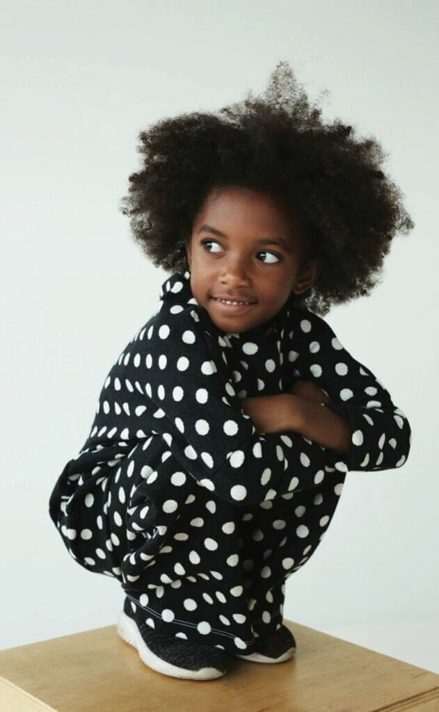 A child in navy polka-dot pyjamas squats on their toes and grins at someone over their shoulder